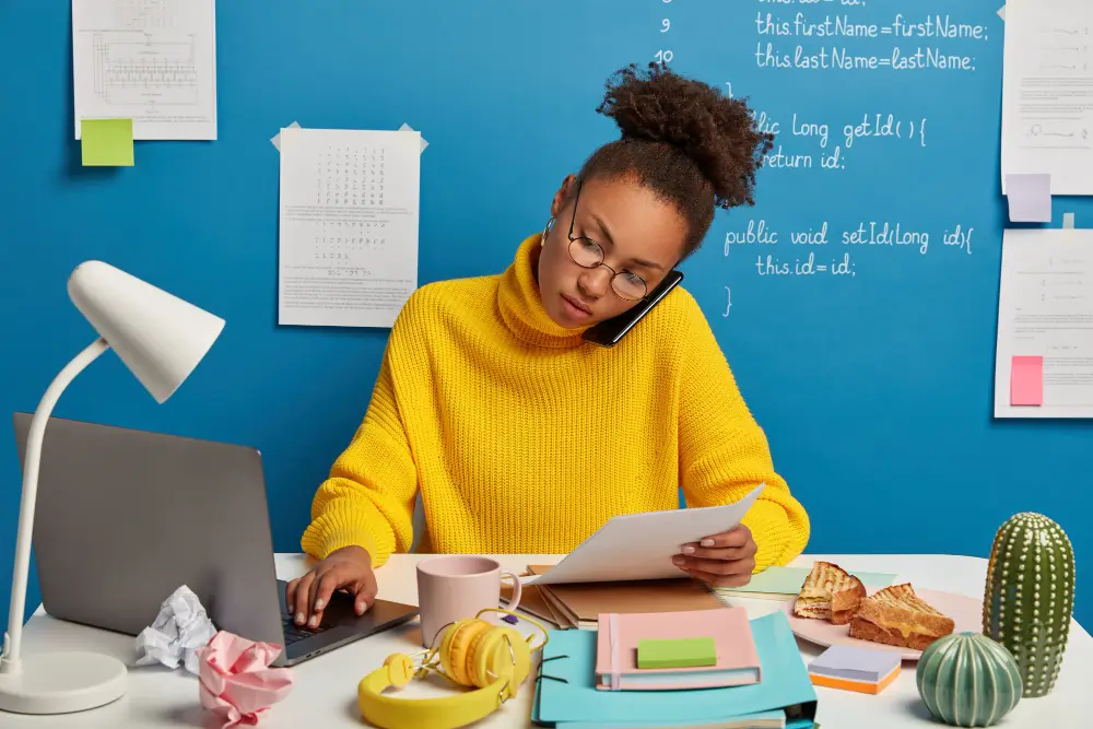 A content writer wearing a yellow turtle-necked shirt, writing on her computer while taking a glance at the documents on her other hand. Their is a cup of coffee on the table, and at the background is a blue board with sticky notes and white chalk draft on it.